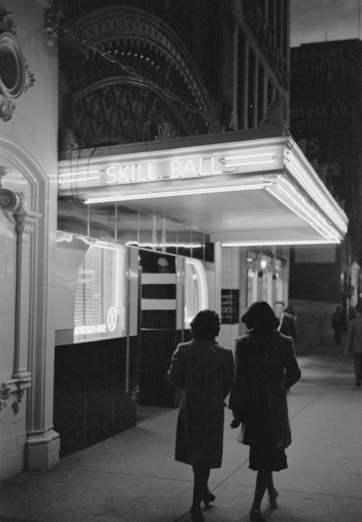 Two women walking under a bright theater marquee on a Butte, Montana street at night, 1939
