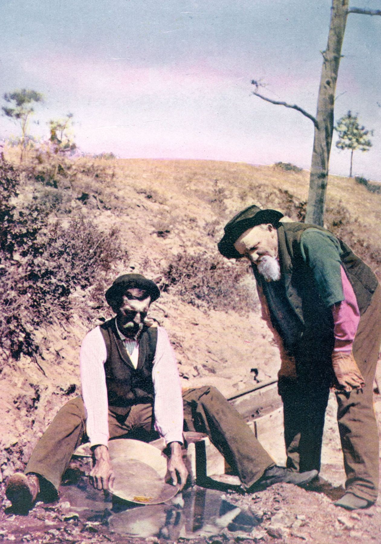 Hand-colored print of two bearded prospectors panning for gold beside a hillside near Butte